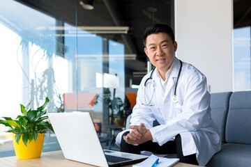Asian doctor is happy and smiling, working in the clinic office on a laptop, looking at the camera - Powered by Adobe
