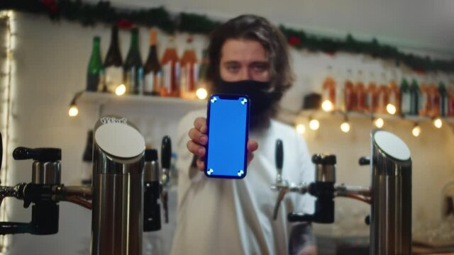 Barman in mask showing blue screen on phone. Bearded man behind bar counter holding smartphone with chroma key. Visiting pubs during coronavirus, green zone restrictions. Bartender in restaurant.
