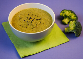 on a lilac background there is a green plate with green broccoli puree soup sprinkled with black...