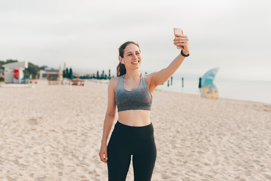 Smiling woman doing sport exercises on morning sunrise beach in sports wear, making selfie photo on phone