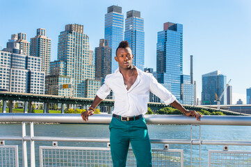 Dressing in a white shirt and green pants,  a young black guy with mohawk haircut is standing in the front of modern high buildings and looking forward..