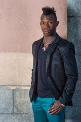 Dressing in a black blazer, green pants, a young black guy with mohawk hair is standing against the wall, confidently looking at you..