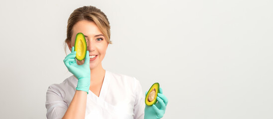 Portrait of young female nutritionist doctor with beautiful smile posing at camera hiding eye behind half avocado on white background, copy space. Benefits of proper nutrition