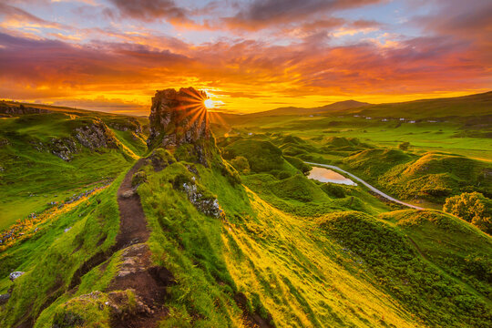 Sunset on the Isle of Skye in Scotland. Sun rays on the mountain hill. Red and orange clouds in the sky. Castle Ewen with paths and meadow in the evening. small lake with road. Sunshine in the valley