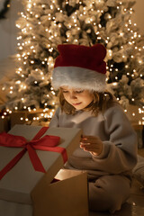 Christmas spirit. Happy little smiling girl opening gift box sitting on the floor near the...