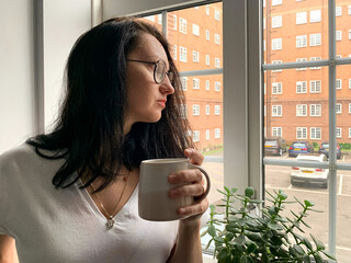 Deep in thought. Young woman is drinking coffee by the window at home. Covid restrictions, self...