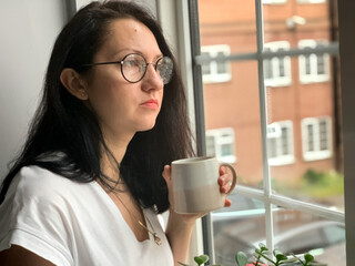 Deep in thought. Young woman is drinking coffee by the window at home. Covid restrictions, self...