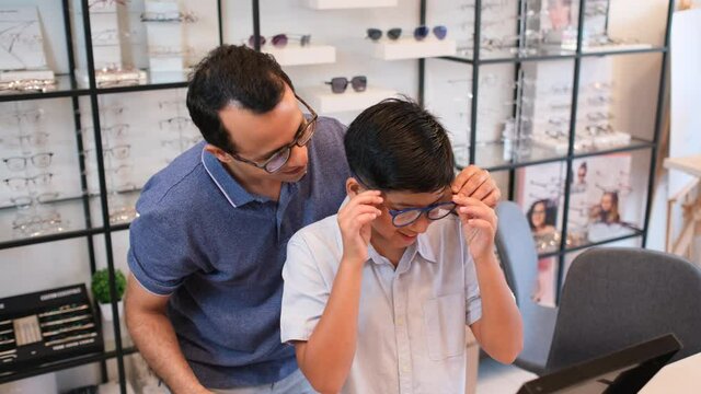 Indian father hold glasses that his son wear and look to mirror with happiness during come to optical shop for buying new glasses.