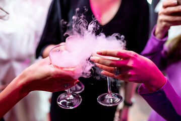 Hands of three girls holding glasses with champagne and dry ice. Beautiful white steam from glasses