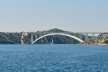 Yachts sail along the gulf of the Adriatic Sea towards the large arched road bridge that connects the islands of Croatia