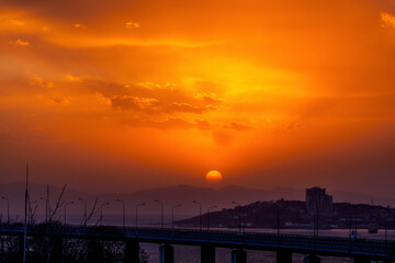 Bright sunset in Vladivostok. The red sun sets on the hills against the background of the road and the sea.