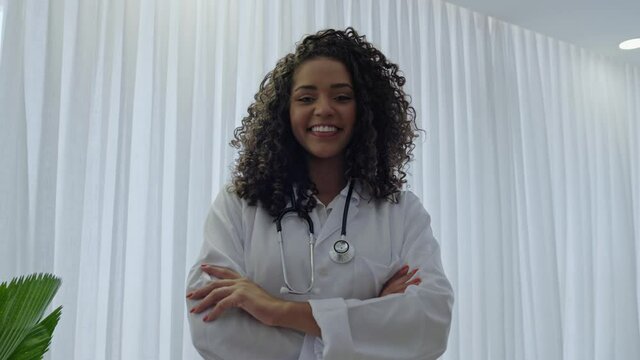 Latin young female doctor wear white uniform, white medical coat, stethoscope and looking at camera in hospital office. Brazilian female doctor, therapist, nurse. 4K.