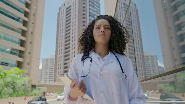 Latin young female doctor wear white uniform, white medical coat, stethoscope and looking at camera in hospital office. Brazilian female doctor, therapist, nurse. 4K.