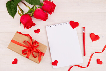 Valentine's Day background. A bouquet of red roses, a gift with a red ribbon, an empty notebook with a place for text and heart-shaped decorations on a white wooden background. Top view