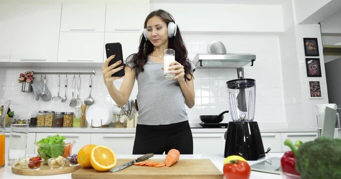 Positive pregnant female shooting selfie video and drinking refreshing milk while looking at camera and dancing in kitchen at home