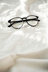 Stylish eyeglasses on white bedding in sunlight with shadows. Flat lay fashion accessories. 