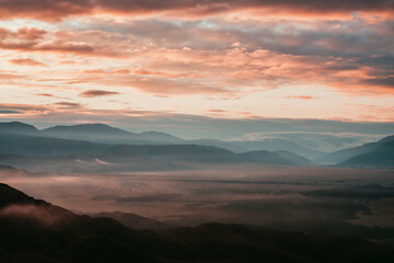 Surreal mountain landscape with pink low clouds above village among mountains silhouettes under dawn cloudy sky. Atmospheric alpine scenery of countryside in low clouds in sundown magenta color.