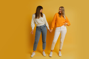 Two pretty women , best friends in stylish autumn  casual clothes having fun  over yellow background in studio. Full lenght.