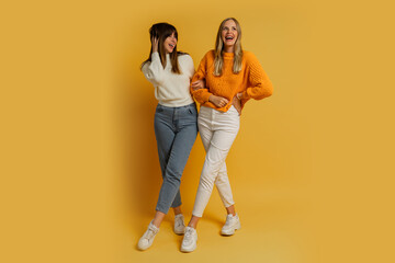 Two pretty women , best friends in stylish autumn  casual clothes having fun  over yellow background in studio. Full lenght.
