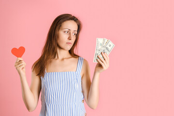 The choice between love and money. A woman holds a heart symbol in her hands and looks at the money in her hand. Studio shot on a pink background
