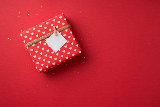 Top View Photo Of Valentine's Day Decorations Shiny Sequins Giftbox In Red Wrapping Paper With Heart Pattern And Twine With Pinned Note On Isolated Red Background With Blank Space