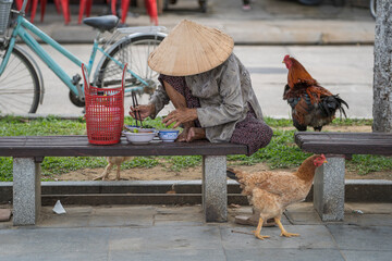 Vietnamese old woman in a straw hat sits on a bench and eats food on a street in the old city in...