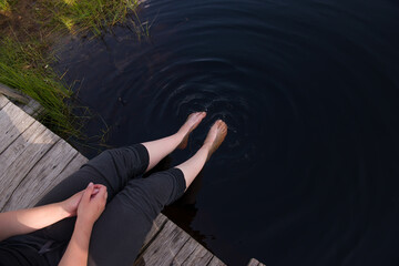 A girl is resting by the lake, sitting on the edge of a wooden pier, her legs dangling along the...