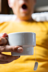 Young man yawning and holding a white teacup. Selective focus.