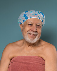 A man with a gray beard aged in a shower hat and a towel, looks and smiling on a blue background, vertical format