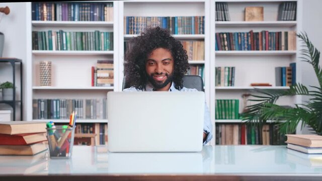 Young inspired curly Indian man freelancer opens laptop and starts working day, having come up with new idea, embodies it at home office sits at large table. People, business, technology concept.