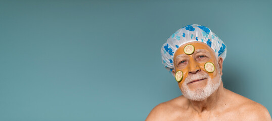 A man with a gray-haired beard aged in a shower cap, with a moisturizing mask of clay for face and sliced cucumbers, concept of relax and body care, on a blue background, banner