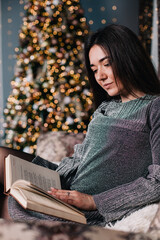 Obraz premium Beautiful sexy woman with Xmas tree in background reading a book sitting on chair. Portrait of a woman reading a book sitting comfortable with a blanket on legs. Attractive brunette female relaxing.