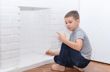 Naklejka premium Child boy sits on the floor and paints a decorative fireplace with white paint gouache. Home improvement, DIY concept. Selective focus