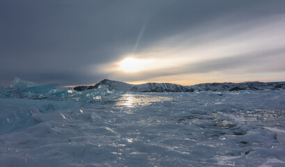 Frozen Lake Baikal in the evening. Reflections of the setting sun on the ice. A block of turquoise shining hummocks against the background of a mountain range and a cloudy sky.