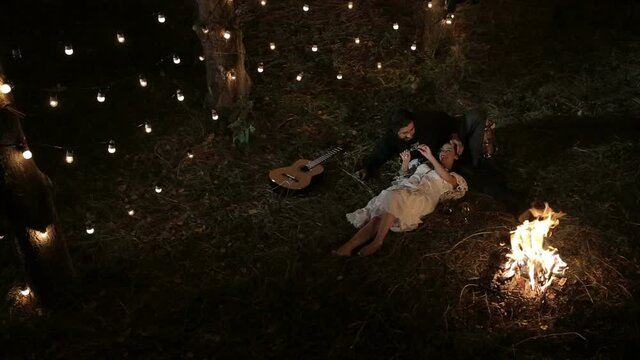 Romantic couple. A brutal guy with a guitar and a girl in a white dress at night in the forest lie in arms on the grass by the fire, drink wine from glasses, talk, laugh and fool around. Garlands 