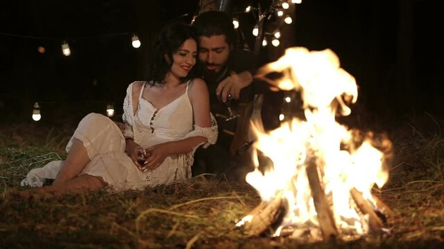 Romantic couple. Brutal guy with a guitar and a girl in a white dress at night in the forest sitting in an embrace on the grass by the bonfire, drink wine from glasses, talk, laugh and fool around. 