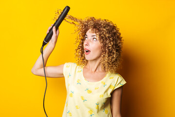 young woman with curly hair straightens her hair with a hair straightener on a yellow background