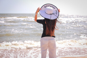 beautiful woman in sunhat white pants, black blouse, standing with her arms raised to her head on summer day.