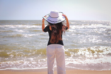 beautiful woman in sunhat white pants, black blouse, standing with her arms raised to her head on summer day.