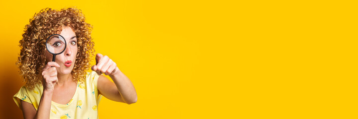 curly girl pointing the finger in front of a looking through a magnifying glass on a yellow...