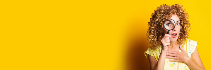 surprised curly girl looks through a magnifying glass on a yellow background. Banner.