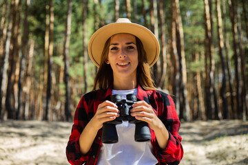 Woman in hat and red plaid shirt holding binoculars in the forest