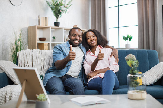 Beautiful and happy couple sitting on couch in embrace and smiling on camera. African american man and woman enjoying leisure time together and showing thumbs up.