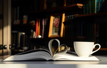 Background - bookshelves lit by the sun. An open book is on the table. The pages are folded in the shape of a heart. On the right is a cup of coffee.