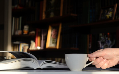 Background - bookshelves lit by the sun. An open book is on the table. Woman's hand with a cup of coffee