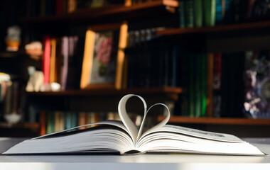 Background - bookshelves lit by the sun. An open book is on the table. Pages are folded in the shape of a heart.  Concept - Valentine's Day