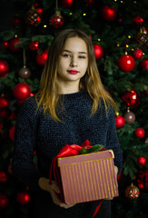 New Year Christmas. Young beautiful blonde girl stands near the Christmas tree decorated with red balls. The girl is dressed in a dark sweater, holding a New Year's gift in a red box.