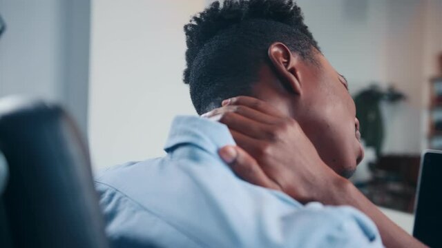 Young African American man office worker grabs neck due to pain suffers from joint injury due to sedentary work tries to stretch sits in office back view. Human health, spine problems concept