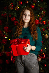 New Year. A young girl with red hair in a green sweater near the Christmas tree, the girl holds a gift in her hands.
