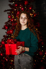 New Year. A young girl with red hair in a green sweater near the Christmas tree, the girl holds a gift in her hands.